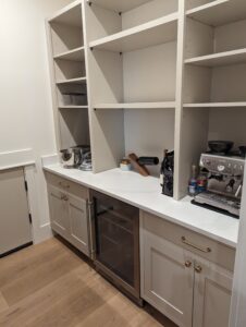 A modern pantry with white shelves and cabinets, a countertop holding a stand mixer, coffee maker, and coffee supplies, plus a mini fridge and pantry items organized neatly.