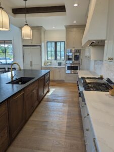 Modern kitchen with light wood floors, an island with a dark countertop and sink, marble countertops, double ovens, large fridge, and windows letting in natural light. Pendant lights hang from the ceiling.
