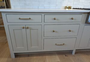 Light gray kitchen cabinet with gold handles and knobs, featuring two drawers on top, a double door on the left, and two larger drawers on the right, set against a tile backsplash and wood floor.