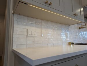White kitchen countertop with light gray cabinets above, under-cabinet lighting, glossy white subway tile backsplash, and three wall switches and one outlet in the center.