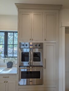 Built-in stainless steel double oven with French doors, set within light-colored cabinetry. A countertop with a potted plant and a window with black trim are visible to the left.