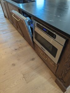 A kitchen with wooden cabinets, one of which has a slanted open panel revealing wiring. Next to it is a built-in Viking microwave oven below a dark countertop. The floor is light wood.