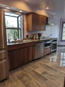 Modern kitchen with wood cabinets, stainless steel appliances, dishwasher, oven, and microwave. Large windows provide natural light. Gray and white patterned tile floor reflects the surroundings. Counter has a knife block and utensil holder.