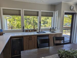 A modern kitchen with wood cabinets, a white countertop, and large windows overlooking green trees. There is a sink under the windows, a dishwasher, a beverage fridge, and a table with a small plant centerpiece.