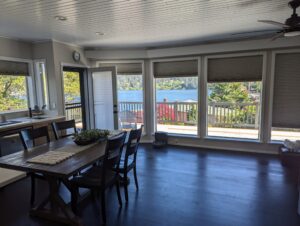A bright dining area with dark wood table and chairs, large windows with shades, and a view of a blue lake and greenery outside. An open door leads to a deck overlooking the scenic outdoors.