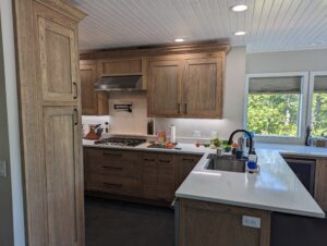 Modern kitchen with wooden cabinets, a gas stove, white countertops, and a sink. Vegetables and utensils are on the counter, and sunlight streams in through windows with a view of green trees outside.