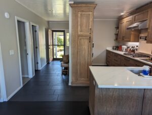 Modern kitchen with wooden cabinets and a white countertop island, next to a hallway with dark tile flooring leading to a glass door with views of greenery outside.