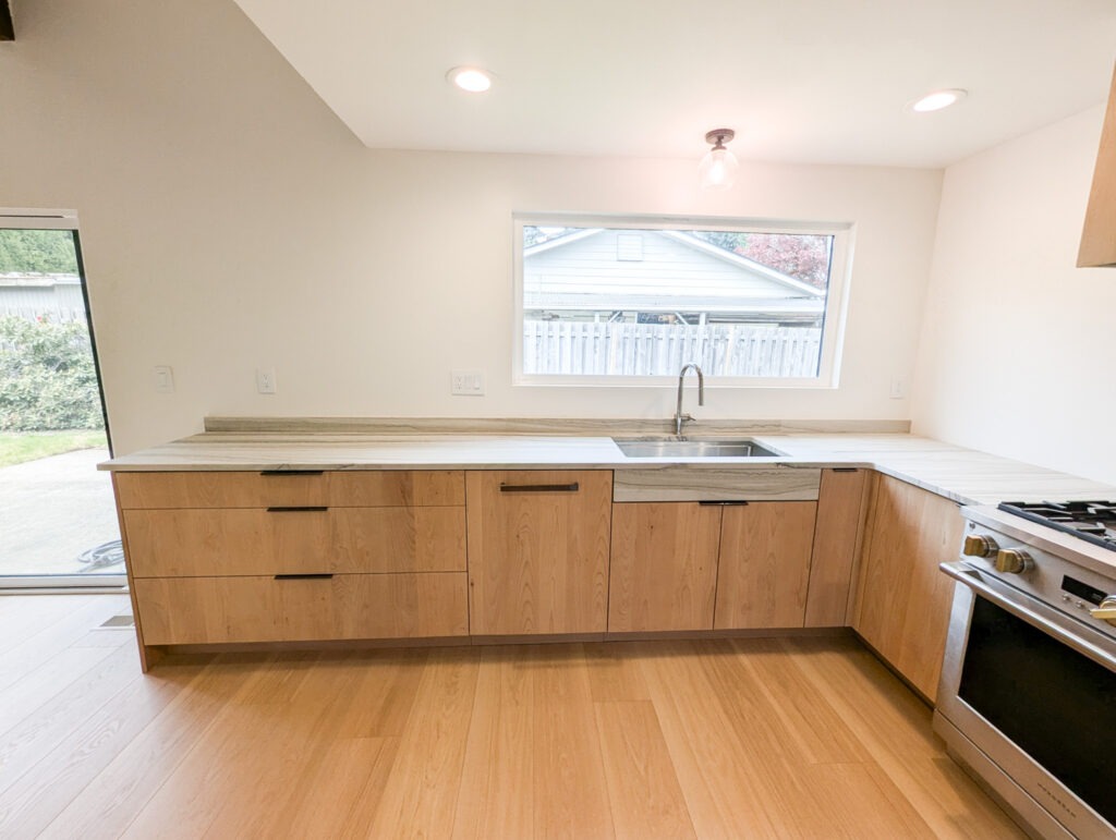 Modern kitchen with light wood cabinets, a stainless steel sink below a large window, and a gas stove on the right. Natural light enters through the window and sliding glass door on the left.