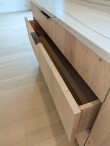 A light wood drawer beneath a marble countertop is open, revealing an empty interior. The cabinet and floor are both made of light-colored wood, and the scene appears to be in a modern, minimal kitchen.