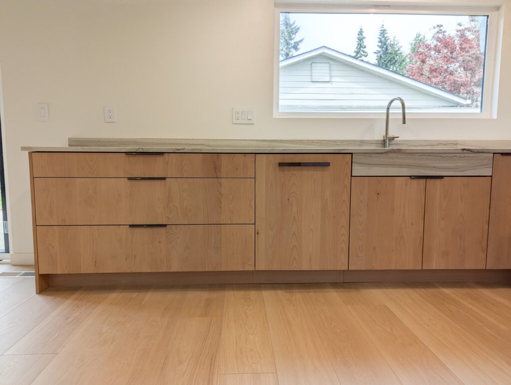 A modern kitchen with light wood cabinets, black handles, a built-in dishwasher, and a light stone countertop. A large window above the sink shows trees and a house outside.