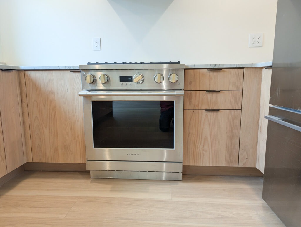 A modern kitchen features a stainless steel oven with four knobs, light wood cabinets and drawers, a marble countertop, and a section of a refrigerator on the right.