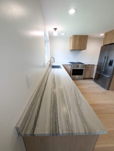 Modern kitchen with light wood cabinets, a stainless steel stove and refrigerator, and a long, marble-patterned countertop with gray veining. A sink is under a window, and the space is well-lit and minimalistic.