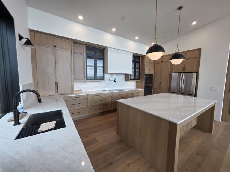 Modern kitchen with light wood cabinets, marble countertops, black faucets, a double-door refrigerator, two pendant lights above an island, and built-in ovens. The space has wood flooring and recessed ceiling lights.