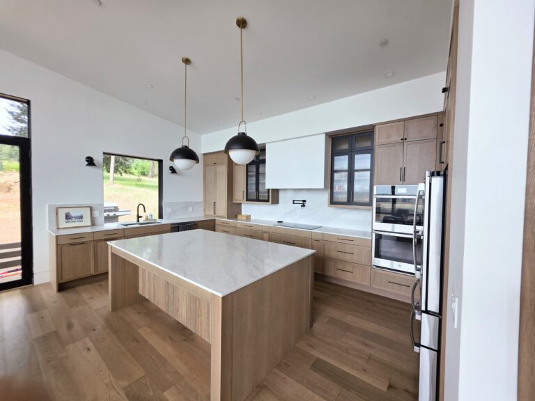 Modern kitchen with light wood cabinets, a large marble island, two pendant lights, stainless steel appliances, and large windows providing natural light.